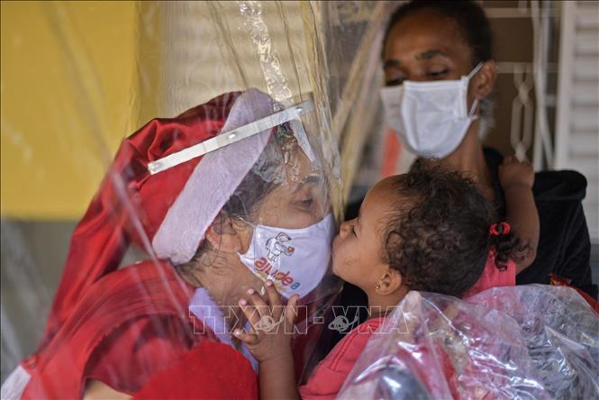 Trong ảnh: Một tình nguyện viên trong trang phục Bà già Noel hôn một em nhỏ qua tấm chắn ni lông nhằm ngăn chặn sự lây lan của dịch COVID-19 tại Minas Gerais, Brazil ngày 7/12/2020. Ảnh: AFP/TTXVN