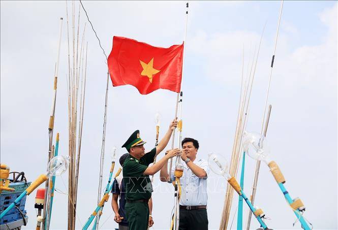 Cán bộ Đồn Biên phòng Sông Đốc (huyện Trần Văn Thời, tỉnh Cà Mau) hỗ trợ ngư dân treo cờ Tổ quốc trên tàu đánh bắt xa bờ. Ảnh: Hồng Đạt - TTXVN
