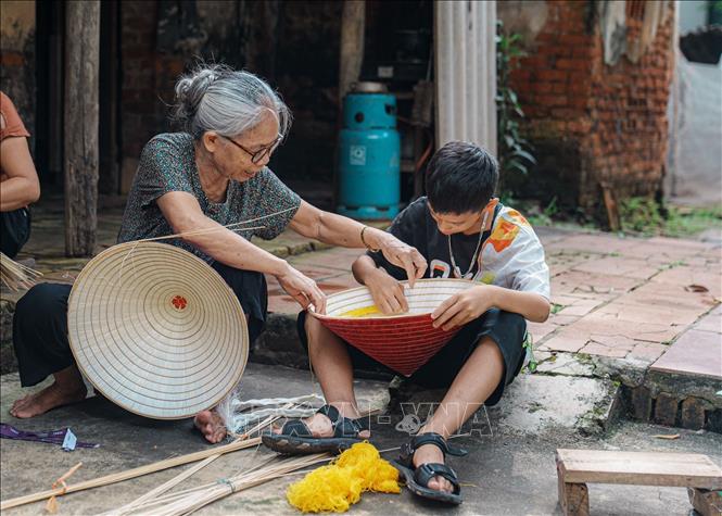Bà Phạm Thị Ba dạy cháu nhỏ cách nhôi nón (trang trí thêm những họa tiết, màu sắc của sợi chỉ khâu để luồn quai nón). Ảnh: Phạm Tuấn Anh-TTXVN