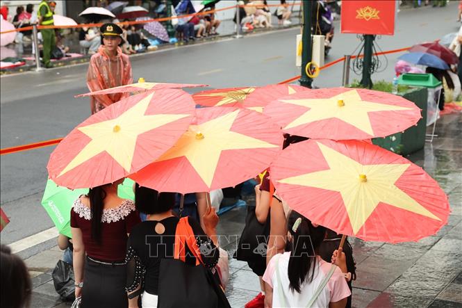 Người dân sử dụng những chiếc ô in hình cờ đỏ sao vàng để che mưa trong lúc chờ đợi buổi Tổng hợp luyện diễu binh, diễu hành lần 2. Ảnh: Vũ Quang – TTXVN