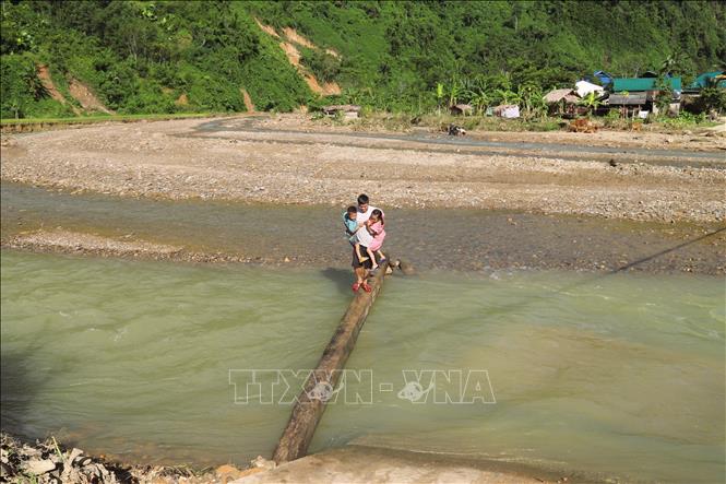 Người lớn bế trẻ nhỏ đi trên thanh gỗ lớn được bắc qua suối Nậm Ngân tại vị trí cầu gỗ đã bị lũ cuốn trôi. Ảnh: Xuân Tiến - TTXVN