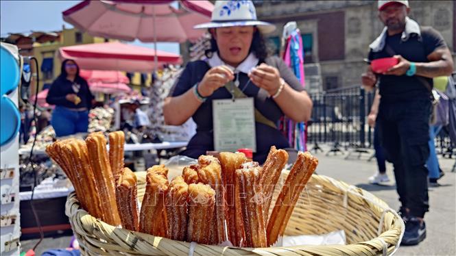 Tiểu thương bán bánh churros truyền thống đón tiếp khách mua hàng trên nền quảng trường Zócalo rợp bóng quốc kỳ Mexico và không khí chuẩn bị lễ hội nhộn nhịp. Ảnh: Phi Hùng - PV TTXVN tại Mexico